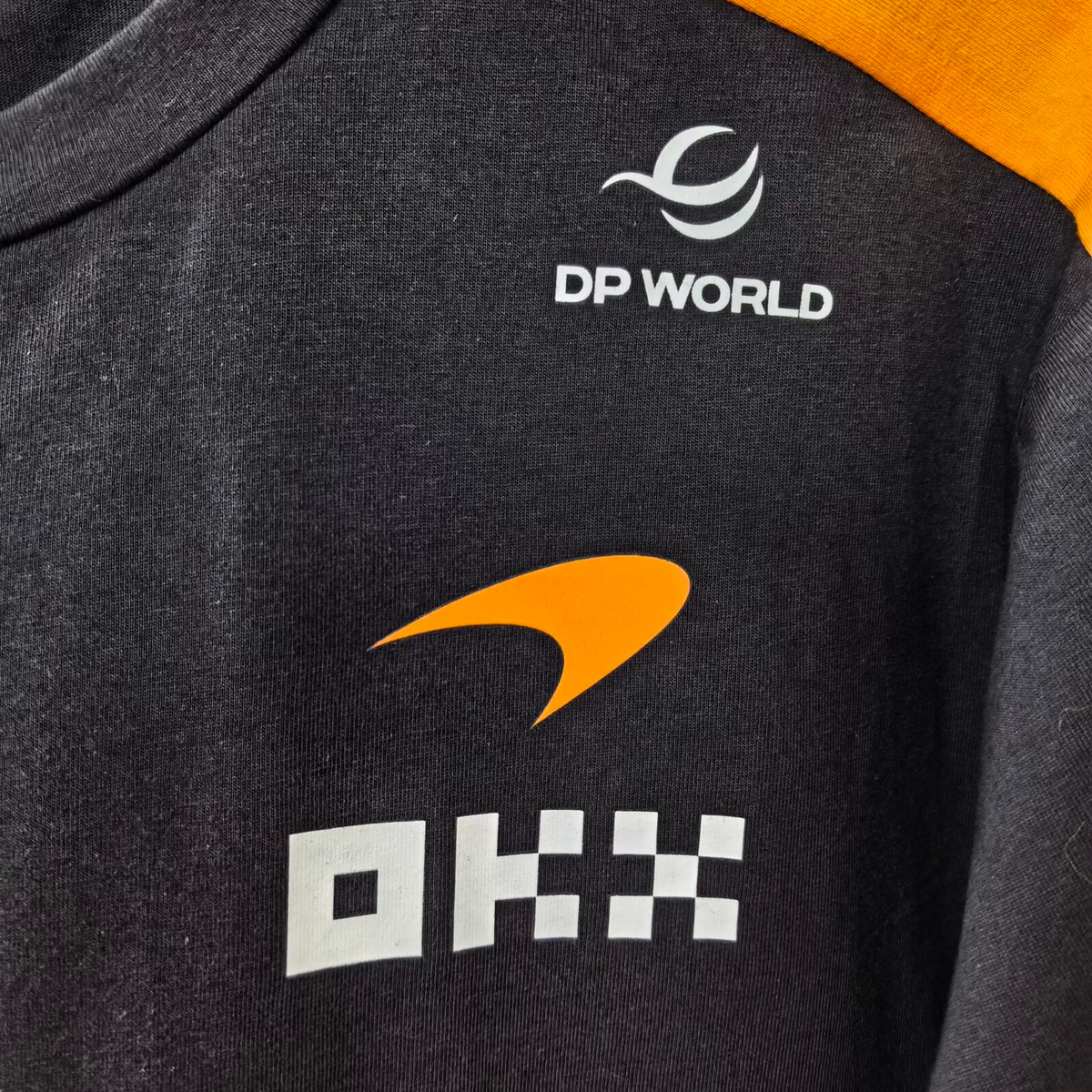 A papaya orange and charcoal McLaren F1 2026 team t-shirt featuring a crew neck and official partner logos including Android, Dell Technologies, OKX, and Optimum Nutrition across the chest.