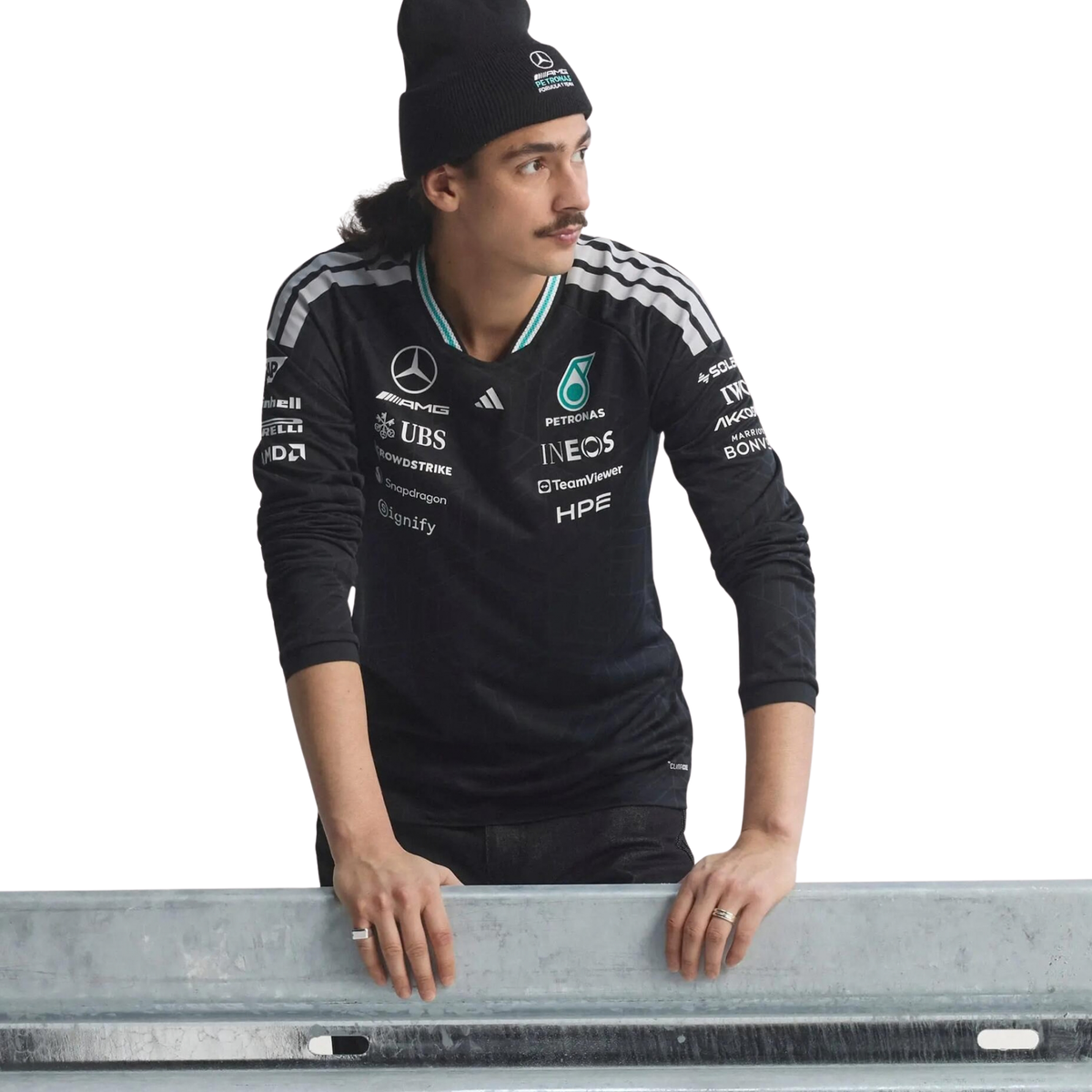 A black Mercedes-AMG Petronas 2026 team long-sleeve t-shirt featuring an all-over tonal geometric pattern, a V-neck collar with white shoulder stripes and vibrant teal inner trim, and official partner logos including Mercedes-AMG, Petronas, and UBS across the chest.