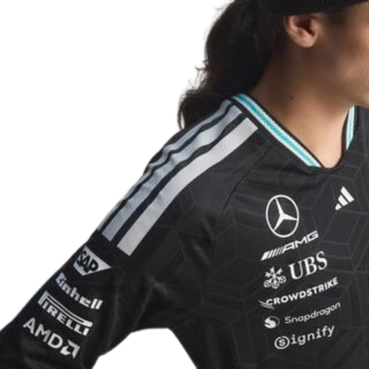 A black Mercedes-AMG Petronas 2026 team long-sleeve t-shirt featuring an all-over tonal geometric pattern, a V-neck collar with white shoulder stripes and vibrant teal inner trim, and official partner logos including Mercedes-AMG, Petronas, and UBS across the chest.