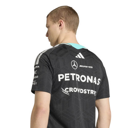 A black Mercedes-AMG Petronas 2026 driver t-shirt featuring an all-over tonal geometric pattern, a V-neck collar with white shoulder stripes and vibrant teal inner trim, and official partner logos including Mercedes-AMG, Petronas, and UBS across the chest.